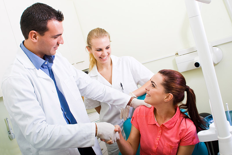 Dentist helping patient in office shaking her hand with assistant in the background
